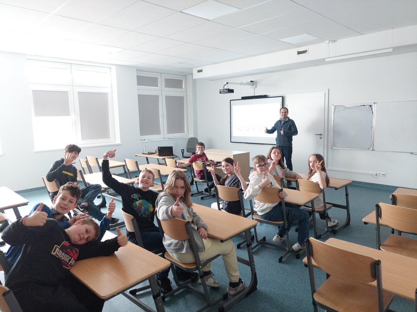 Dzieci i nauczyciel w klasie pokazują dłońmi znak, że jest dobrze, kciuki do góry.