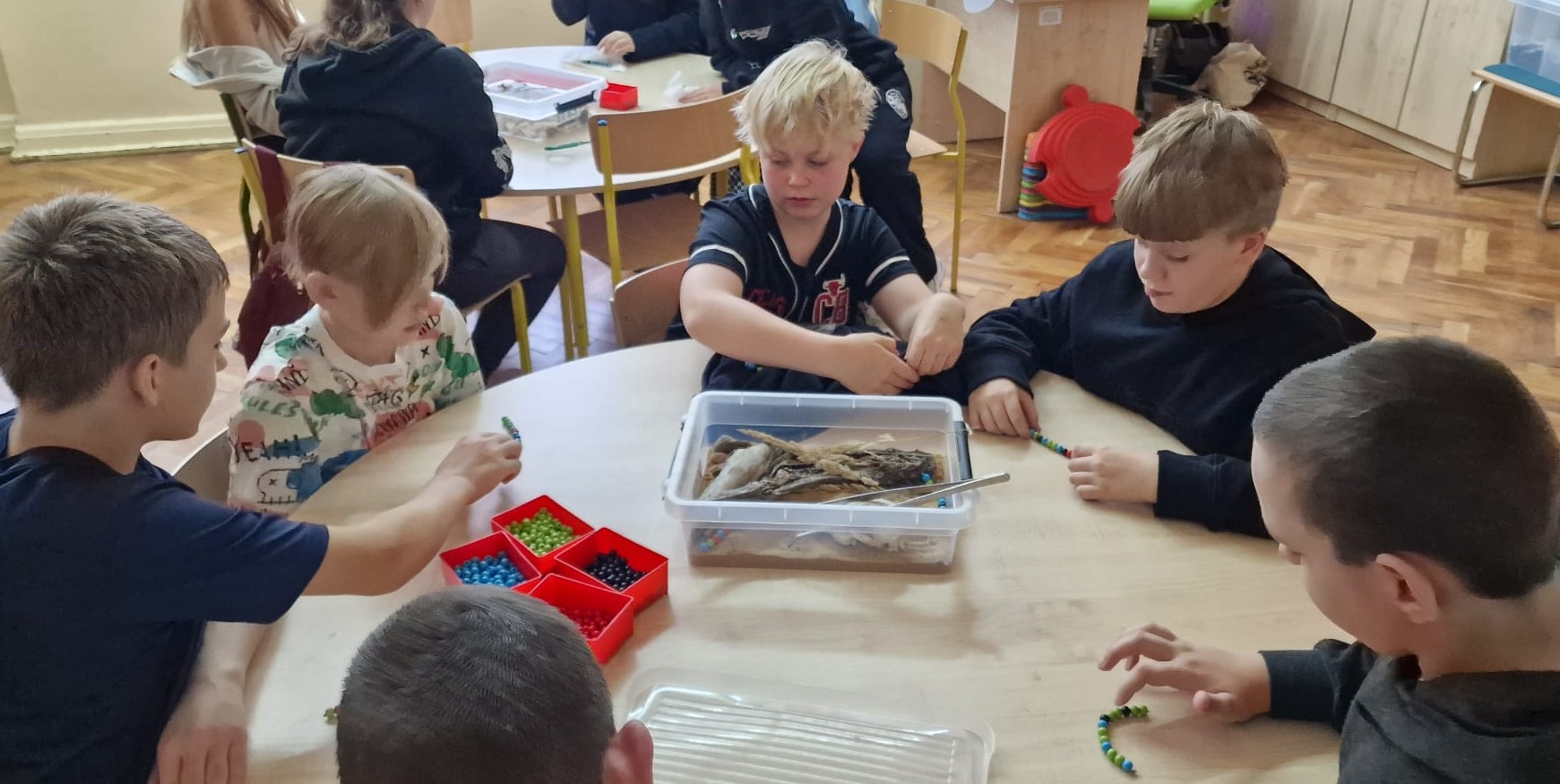 Dzieci w klasie siedzą przy okrągłym stole wykonując ćwiczenie z warsztatów biologicznych.