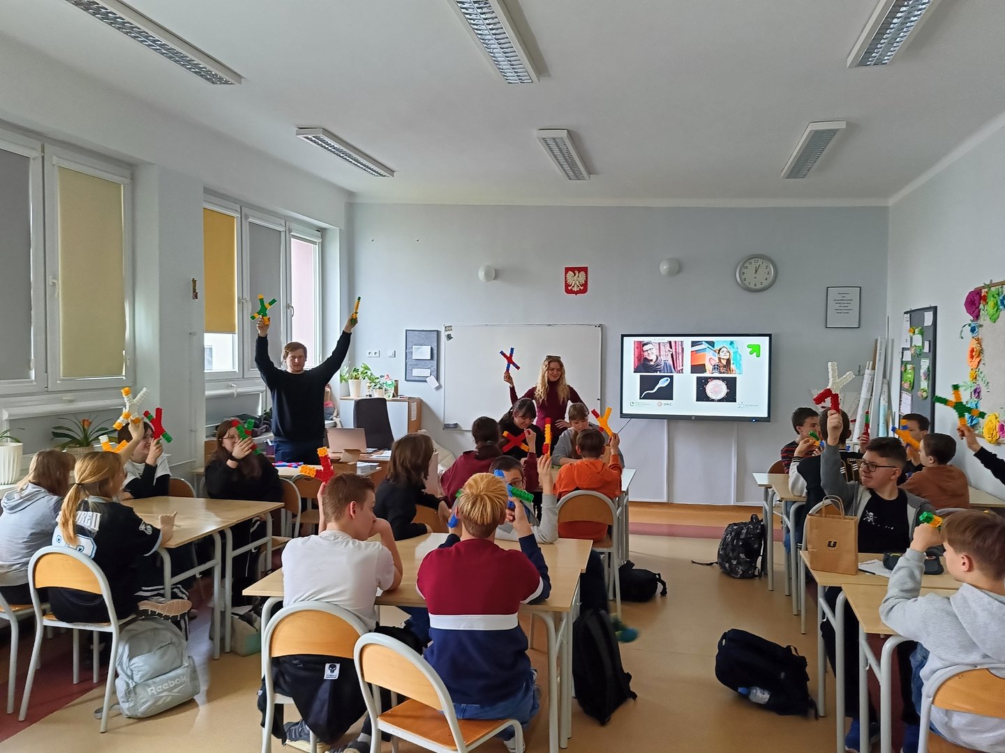 Prowadzący zajęcia trzymają w dłoniach chromosomy z klocków pokazując klasie jak wykonać zadanie.