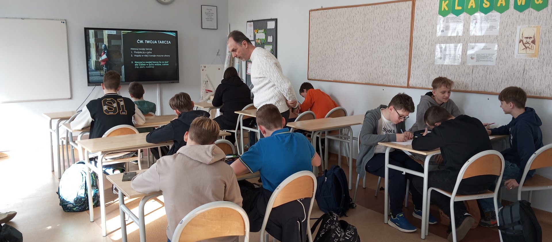 Dzieci siedzą w ławkach i wykonują ćwiczenie pod nadzorem nauczyciela